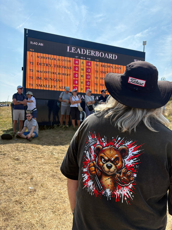 A person standing outdoors in a field wearing a black T-shirt with a large graphic of a bear holding a gun and a black hat. A large leaderboard scoreboard is visible in the background with several people around it under a blue sky.