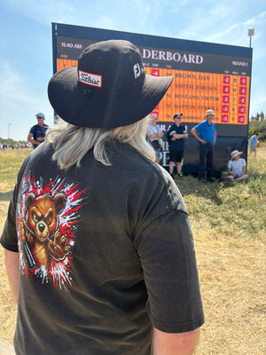 A person standing outdoors at a golf event, viewed from behind, wearing a black wide-brim hat with a Titleist logo and a dark gray T-shirt with a large bear graphic on the back. A large leaderboard scoreboard is visible in the background under a blue sky.