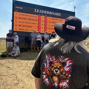 A person standing outdoors in a field wearing a black T-shirt with a large graphic of a bear holding a gun and a black hat. A large leaderboard scoreboard is visible in the background with several people around it under a blue sky.