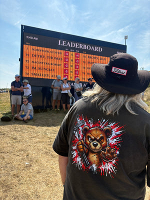 A person standing outdoors in a field wearing a black T-shirt with a large graphic of a bear holding a gun and a black hat. A large leaderboard scoreboard is visible in the background with several people around it under a blue sky.