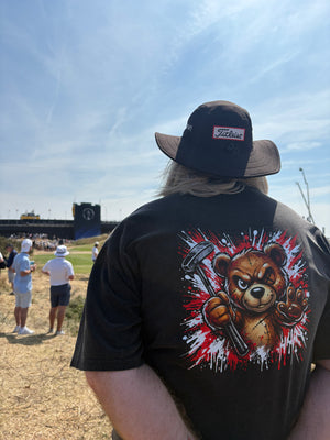 A person viewed from behind wearing a black T-shirt with a large graphic of a bear holding a golf club along with a dark wide-brim hat, standing outdoors on a sunny day at The Open Golf Championship