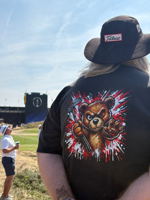 A person viewed from behind wearing a black T-shirt with a large graphic of a bear holding a golf club along with a dark wide-brim hat, standing outdoors on a sunny day at The Open Golf Championship