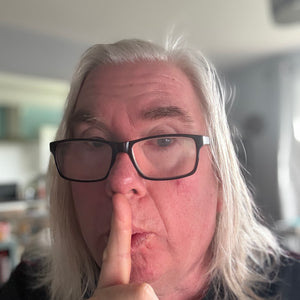 Person called john ogden, big oggie, with white hair and glasses making a 'shh' gesture indoors.