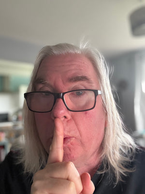 Person called john ogden, big oggie, with white hair and glasses making a 'shh' gesture indoors.