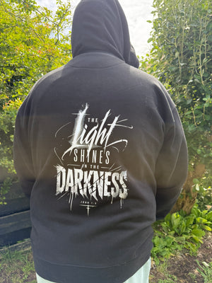 Person wearing a black hoodie with 'The Light Shines in the Darkness' text on the back, standing outdoors.
