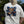 A person viewed from behind wearing a white short-sleeve T-shirt outdoors. The back of the shirt features a large graphic print of a brown teddy bear surrounded by a blue and black paint splatter design.