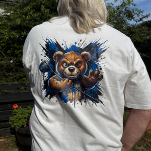 A person viewed from behind wearing a white short-sleeve T-shirt outdoors. The back of the shirt features a large graphic print of a brown teddy bear surrounded by a blue and black paint splatter design.