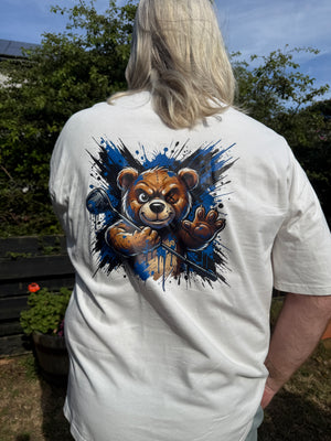 A person viewed from behind wearing a white short-sleeve T-shirt outdoors. The back of the shirt features a large graphic print of a brown teddy bear surrounded by a blue and black paint splatter design.