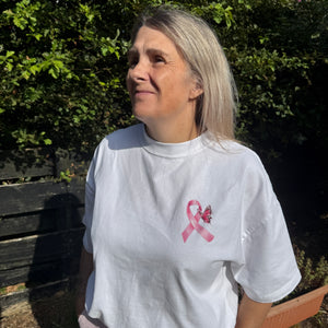 A woman wearing a white t-shirt with a pink ribbon and BUTTERFLY graphic on the chest, photographed outdoors against a background of green foliage and a wooden bench.
