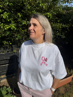 A woman wearing a white t-shirt with a pink ribbon and BUTTERFLY graphic on the chest, photographed outdoors against a background of green foliage and a wooden bench.