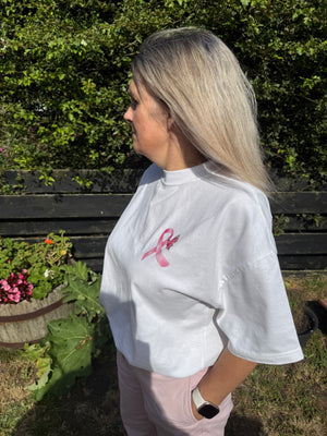 A woman with blonde hair stands outdoors in profile, wearing an oversized white t-shirt with a pink ribbon graphic on the chest and light pink pants.